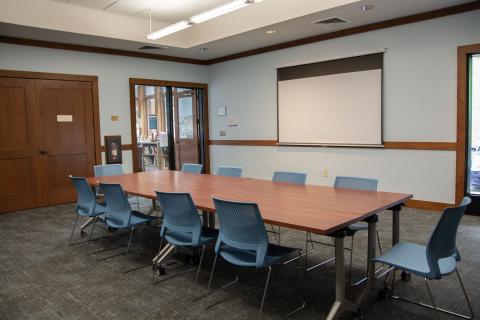 carpeted room with four tables put together to form one big meeting table with 10 chairs around it and one projector screen