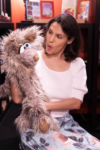 Dark-haired woman talking to a large furry brown puppet with huge eyes.