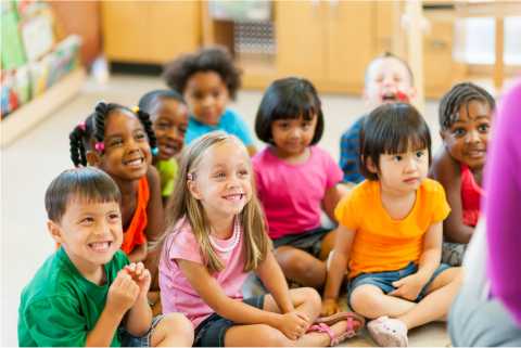 Nine racially and gender diverse, preschool-age children, sitting on the floor in a clump, attentively facing an adult, who is sitting on the floor facing them. We can see only the back of their left arm.