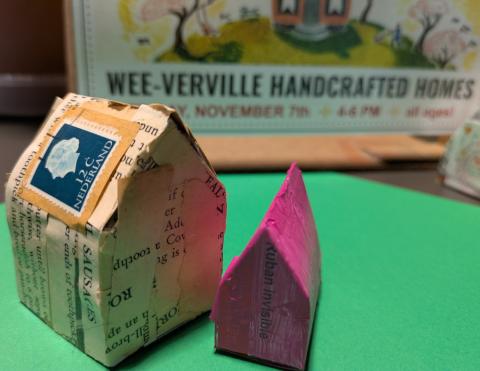 Two tiny handmade paper houses. One is covered with newsprint has a postage stamp on it's tiny roof. The other smaller skinnier house is painted hot pink. Both houses sit on a green paper in front of a decorative sign announcing, "Wee-verville Handcrafted Homes."