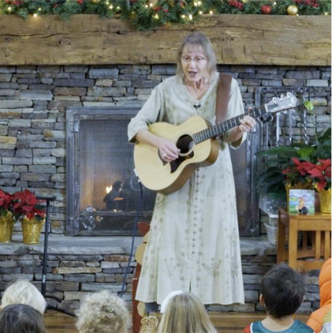 Blond woman with a guitar sings in front of a fireplace with Christmas lights and poinsettias.